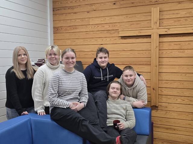 Kuusi nuorta sohvalla, taustalla seinän kokoinen risti
