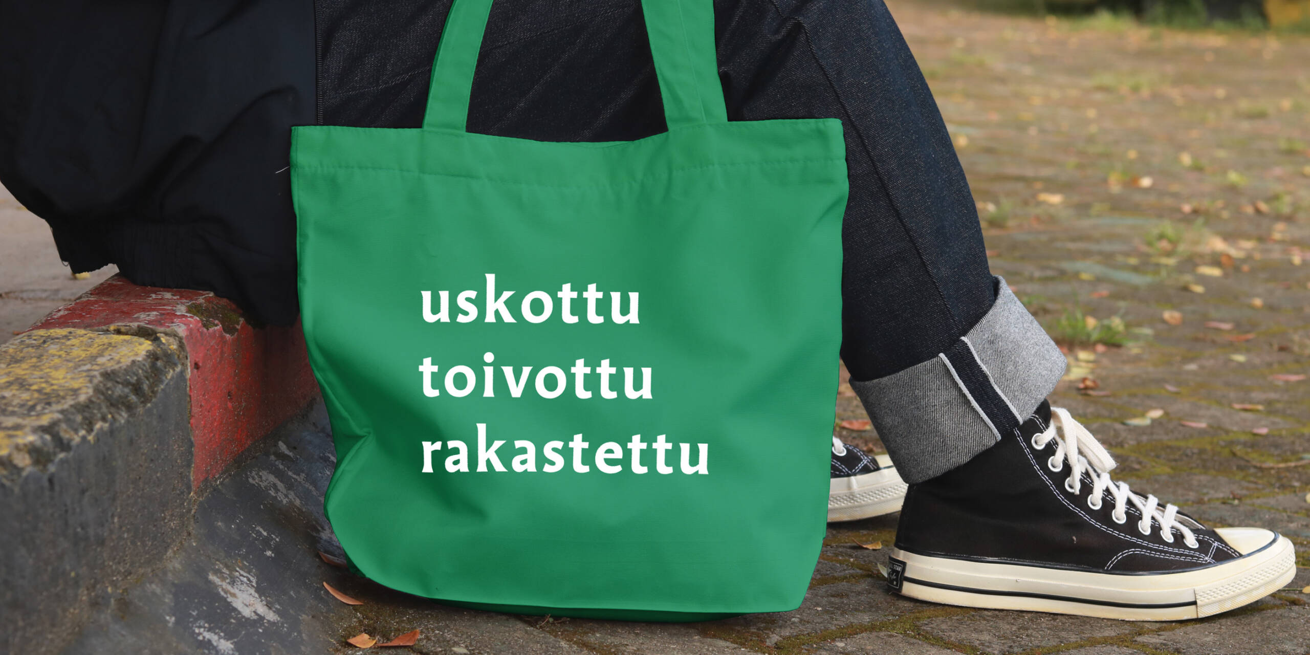 Vihreä kangaskassi, jossa on valkoiset sanat "uskottu", "toivottu" ja "rakastettu". Taustalla henkilö istuu kivetyksellä.