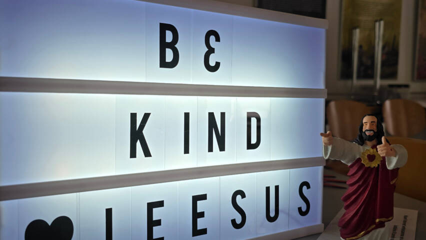 Pöydällä päällä oleva valotaulu, jossa lukee be kind sydän Jeesus. Pöydällä on myös hymyilevä Jeesus-patsas, joka osoittaa valotauluun ja näyttää peukkua.
