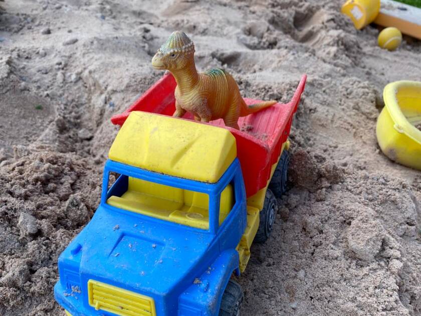Hiekalla monivärinen lelukuormaauto, jonka kyydissä leludinosaurus. Taustalla muita hiekkaleluja ja vähän vihreää nurmikkoa.